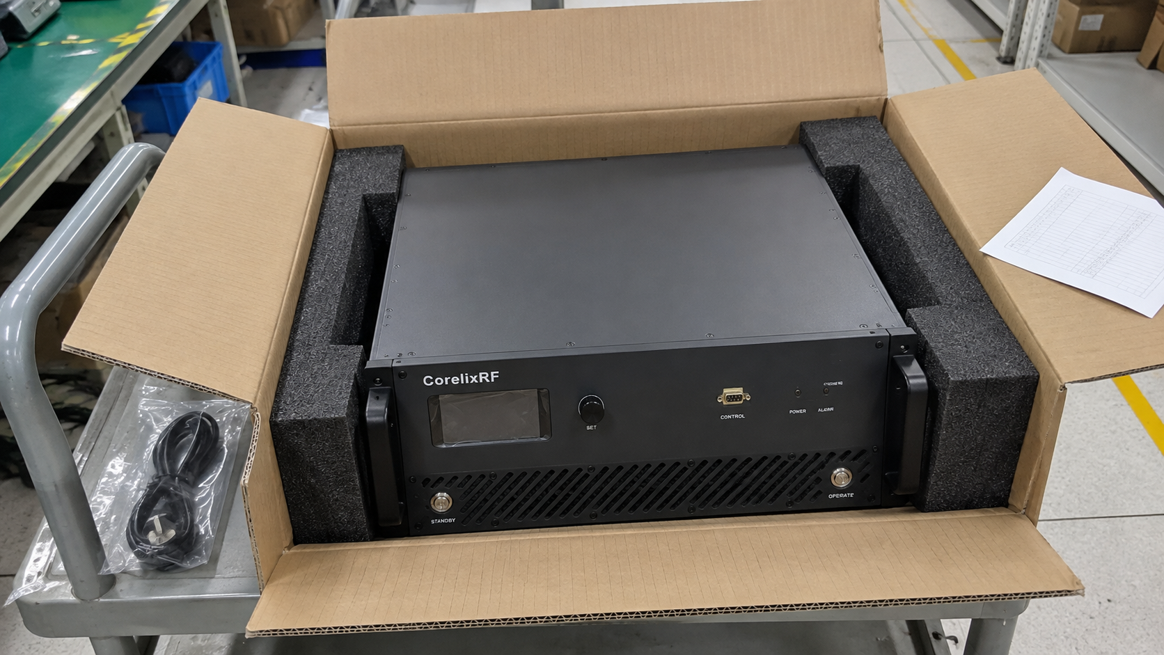 A CorelixRF rack amplifier in protective foam with an accessory pouch at a factory packing station.