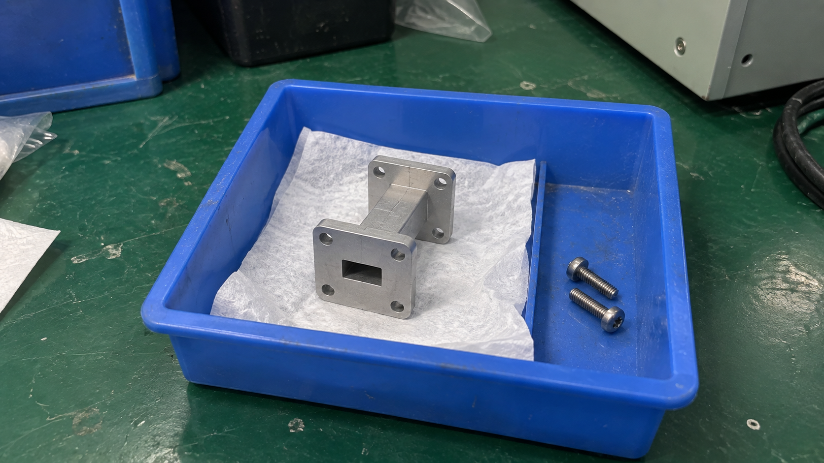 A small rectangular waveguide section resting in a parts tray on a factory inspection bench.