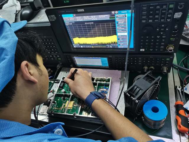 Technician adjusting an open amplifier assembly beside RF test instruments and cooling fans.
